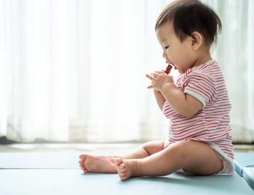 Foto com um bebê sentado levando um item pequeno com as mãos até a boca.