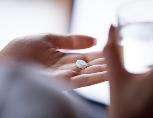 Pessoa com um copo de água na mão direita e um comprimido de nimesulida na mão esquerda, preparando-se para tomar o medicamento.