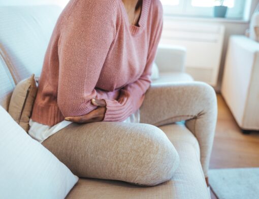 Pessoa sentada no sofá, com as mãos no abdômen, indicando desconforto causado pela síndrome da tensão pré-menstrual.