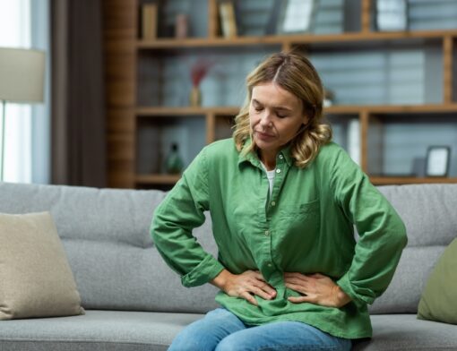 Uma pessoa sentada no sofá de uma sala, apresenta semblante de desconforto e está com as duas mãos pressionando o abdômen.