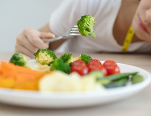 Pessoa sentada à mesa com um prato com uma porção de alimentos e fixamente olhando para um vegetal que acabou de pegar com um talher.