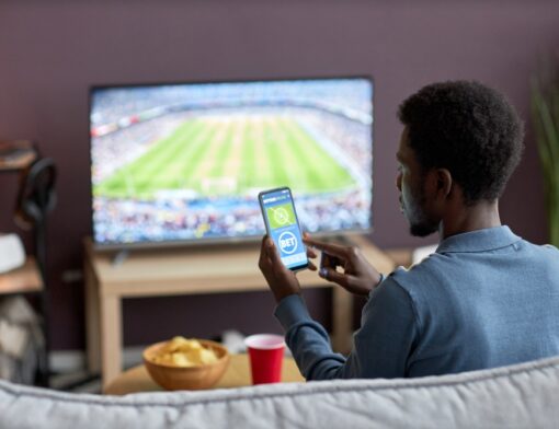 Pessoa assistindo a um evento esportivo em uma sala, está sentada em um sofá com um aparelho de celular nas mãos, acessando um site ou aplicativo de apostas - Bet.