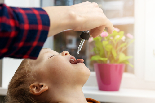 A imagem mostra uma criança recebendo suplementação de vitamina D por meio de gotas administradas com um conta-gotas. Uma mão adulta segura o instrumento enquanto a criança está com a cabeça inclinada para trás e a boca aberta. 