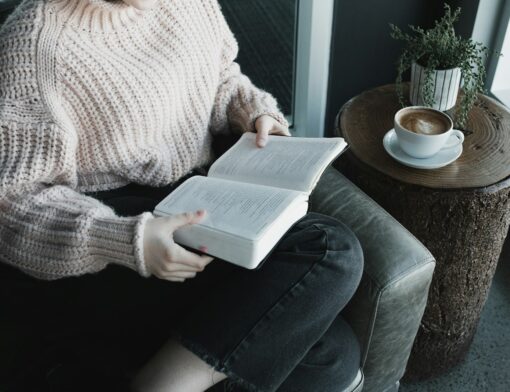 Pessoa sentada em uma poltrona, lendo um livro enquanto saboreia um café.