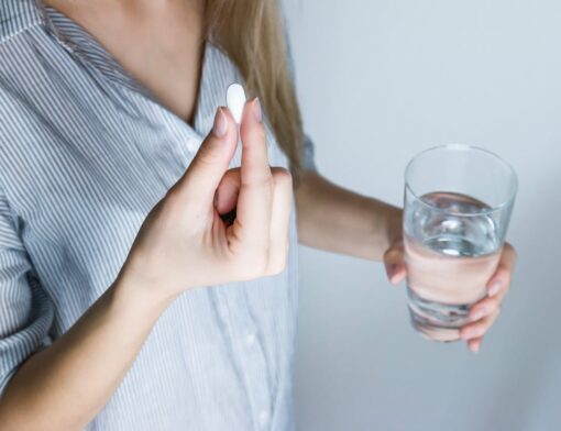 Pessoa com um comprimido na mão direita e, na mão esquerda, um copo com água para se automedicar.