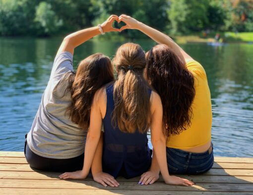 Três jovens sentados em um píer comemorando a amizade com abraços e sinais de coração feitos com as mãos.